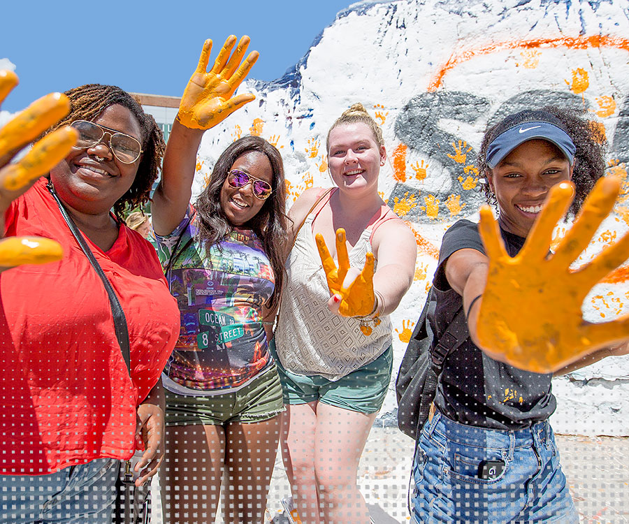 Four young women stand in front of The Rock displaying hands painted orange