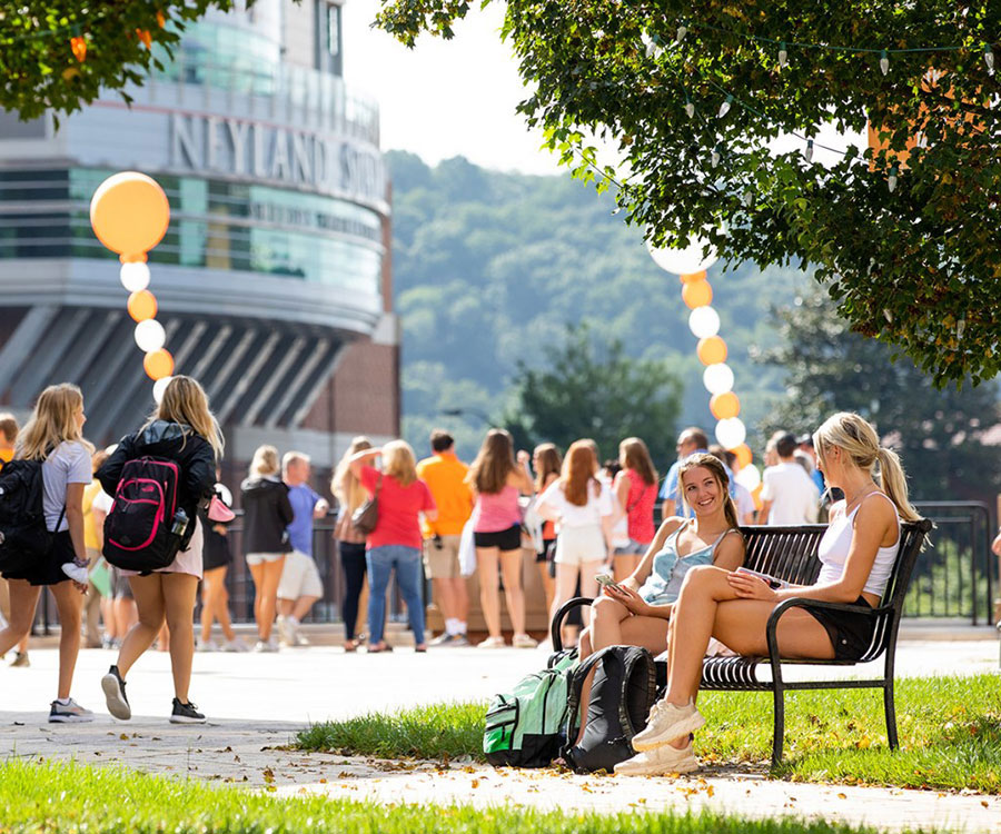 Two UT students sit smiling and talking on a bench with Neyland Stadium and columns of orange and white balloons behind them.