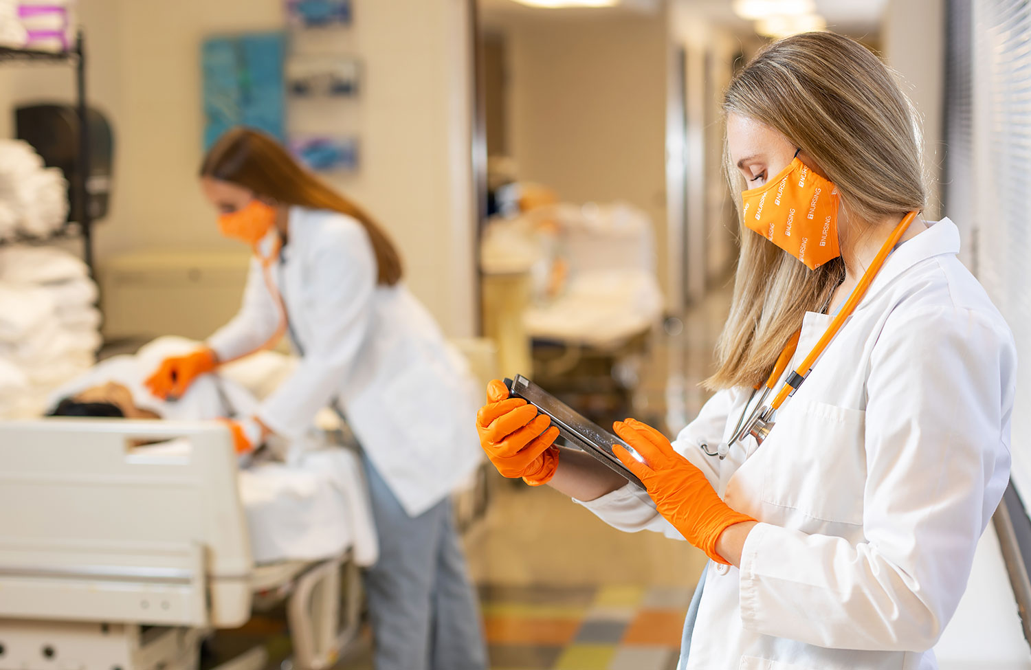 Nursing student wearing white coat, orange face covering and gloves, looks at the V-Visit Sim app on her tablet